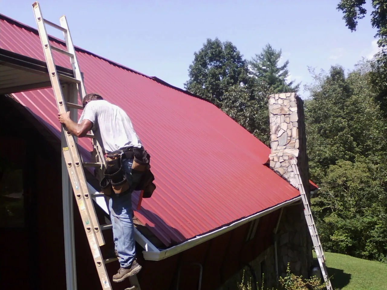 Roofer on a ladder beside a red metal roof and stone chimney — Soffit & Fascia Repair by Roofing Brothers in Ravensdale